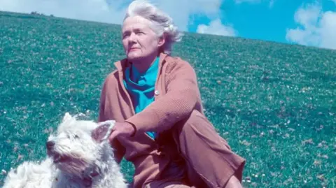 Du Maurier sitting in long grass with her white scottie dog. She wears a smock, blue blouse ands brown cardigan, trousers and desert boots