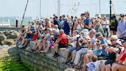 Cowes Week Martin Allen Spectators seated and standing along a harbour wall watching activity on the water during Cowes Week.