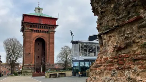 A large, red brick water tower stands in a public square. It has a green roof and a rust-coloured section at the top. Also pictured is the side of a Roman wall, and a modern glass building which houses the Mercury Theatre.