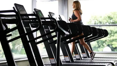 PA Media A stock image of a woman running on a treadmill. She is in a gym with floor to ceiling windows in front of her and next to her. She is running on one of six treadmills lined up and the others are empty.
