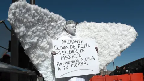 A member of a religious group dressed as an angel takes part in a demonstration at the Ciudad Juarez international crossing from Mexico to the US. They hold up a sign written in Spanish protesting against the the immigration policies of Donald Trump. It reads "Migrants, the God in the US is the God in Mexico. Welcome back home to your land."