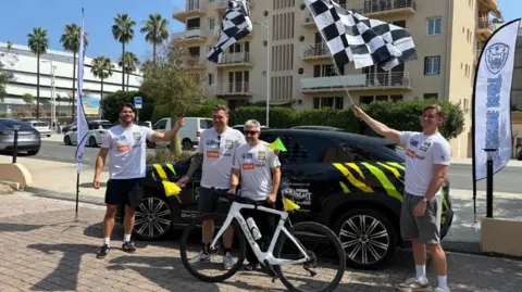 Michael Mansell Four men stand next to a black car in Nice, some waving chequered flags and one standing next to a bike.