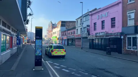 A police car is parked outside a pink building called Pink. Other business premises can be seen on either side of the road. Some smoke from a burning building can be seen in the distance. 