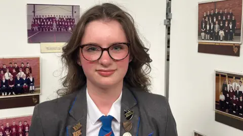 John Devine/BBC Maria has long brown hair and is wearing black framed glasses, a white shirt, a grey blazer and blue tie. She has several badges on her blazer.
