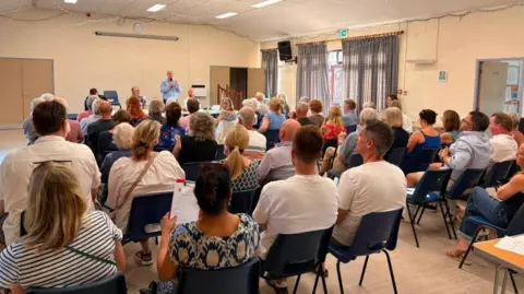 Ben Goldsborough A man stands up holding a microphone in a village hall to address a group of about 50 people who have attended a community meeting, with guests sat on blue chairs.