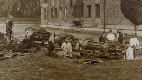 National Trust A sepia-coloured photo from 1910 featuring seven workers standing in and around the moat at Oxburgh Hall. They are wearing hats and some have spades. There are carts on wheels full of excavated earth.