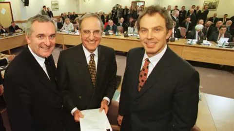 PA Media Three men stand in front of a table in a room full of other politicians. They are all smiling, Bertie Ahern is on the far left. He has short, grey, receding hair. In the middle is George Mitchell. He also has short grey hair and is balding. He is wearing glasses and is holding a big sheaf of papers. Tony Blair is on the right of the picture. He has short brown hair. All three are wearing dark coloured suits with white shirts and multi-coloured ties.