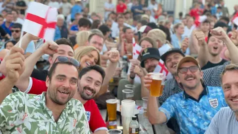 A crowd of people holding England flags and pints of beer are cheering and smiling
