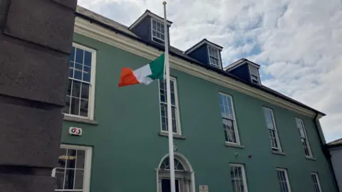 Irish tricolour flying at half-mast on a white pole at Dublin Metropolitan Regional Headquarters at Dublin Castle - the building behind the flag is green