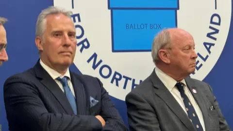 A sombre looking Ian PAisley stands on the elction platofrom, wearing a dark suit and with arms folded. To his right is Jim Allister, looking away from camera. Both mean are standing in front of an electoral office banner