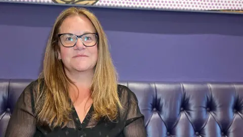 Claire sits on a deep purple, button‑backed bench with bright pop‑art style artwork featuring a crab displayed on the wall above. She is wearing a black, semi‑sheer top and have long, light brown hair resting over their shoulders.