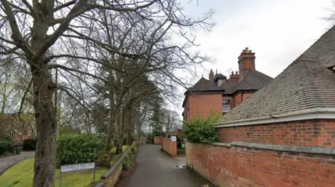 Buildings are visible on the right and far left of the photo. Trees, a bush and some grass are on the left and in the middle.