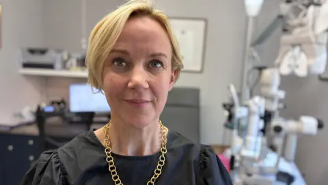 A woman with blonde hair in a short bob. She is wearing a black top and a gold chain necklace. She is in a room in an optician surgery.