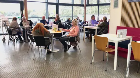 About 10 people sitting at tables in a cafe, with a large window behind them