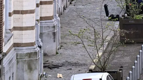 Getty Images The scene of an explosion at the synagogue in the rue Leon Fredericq, in Liege on Monday. It shows a cobbled street with two planters with trees in them. Scorch marks and debris from the blast can be seen on the cobbles.