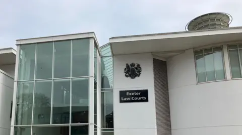 The outside of a large white painted building. It has a big glass window and large entrance area and shows the Exeter Crown Court logo and a plaque that reads Exeter Law Courts. 