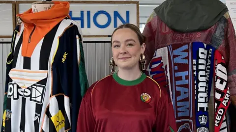 Liv Ashton is wearing a maroon football shirt with green trim round the neck and a club logo on one side of the chest. Behind her are two of her fashion creations – both made out of old football kit but looking like dresses or long coats. The back of one appears to be made from vertical football scarves sewn together.
