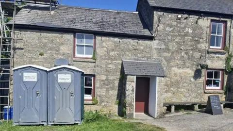 Two portable toilets in the back garden of a pub - Close to the door there are blackboards advertising beer and food offers. 
