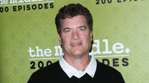 Getty Images Mid shot of Pat Finn wearing a dark sweater and button-up white shirt in front of a green wallpaper backdrop ahead of a viewing of the Middle.
