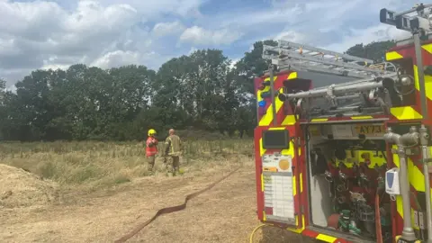 Norfolk Fire and Rescue Service Two firefighters stand in a field alongside a fire engine with a hose unfurled on the ground.