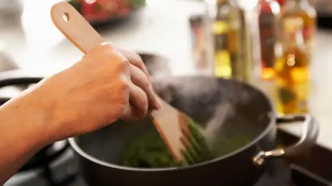 A person stirs a green substance in a black pot with a wooden spatula.  A kitchen counter and condiments are visible in the background but heavily blurred. 