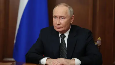 Russian President Vladimir Putin is seated and stares to the left of the camera, wearing a dark suit. 