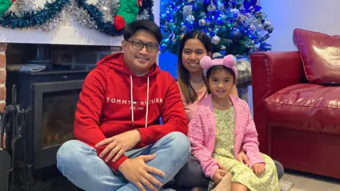 Oliver and Russel Fabia with their daughter. They are sat on the floor in front of a log burner and a Christmas tree.