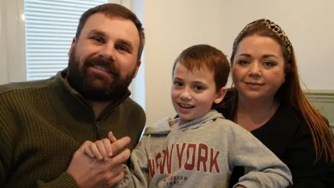 A family look forward at the camera. A man on the left is wearing a dark green jumper. A boy, in the centre, is wearing a grey hooded jumper with the words New York on the front. A woman, to the right, is wearing an animal-print headband in her long dark hair.