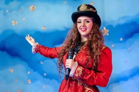 Circus Starr A woman with long curly brown hair performs on a stage. She wears a red coat and a black hat, while holding a microphone. She stands in front of a light blue cloud backdrop with gold stars on it.