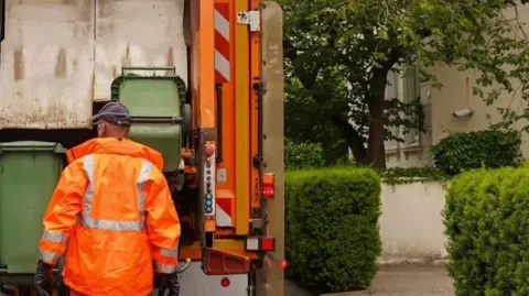 A refuse collector wearing an orange jacket, standing in front of a bin lorry with his back to the camera. To his right is a low hedge, a tree and a just-glimpsed house.