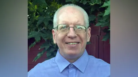 Patrick Mahon A close-up image of a man with white hair smiling at the camera. He wears a light blue shirt and gold glasses.