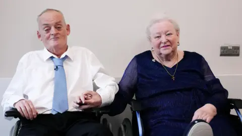 Colin and Pamela sat in wheelchairs holding hands. They are both looking at the camera. Colin, on the left, wears a white shirt, blue tie and black trousers. Pamela, on the right, wears a navy long sleeved dress and a gold necklace. She has short white hair. Pamela has one leg elevated.