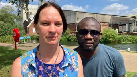 BBC/Sam Read A woman is sat on the left dressed in a summery blue top, a man is behind her on the right wearing sunglasses. The river is behind them, water safety equipment can be see alongside it.