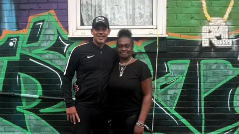 BBC Vincent Nembhard, left, stands next to Sandra Nembhard. They both are wearing black and have one arm wrapped around each other. They are standing in front of a mural on their garden wall which says the word  'stretch' in black and green graffiti font on the bottom.