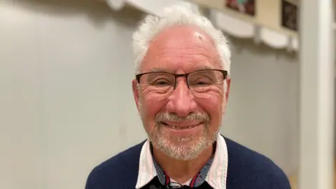 Carl Pearce is looking directly into camera and is smiling. He has white hair, a beard and glasses.