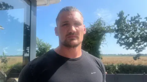 A man stands outdoors near a glass window, dressed in a black 'Paul Smith' T-shirt. Behind him, a scenic landscape unfolds with lush greenery, including trees and bushes, leading to an open field beneath a clear blue sky.