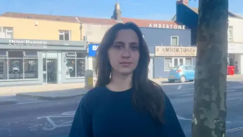 A young woman with dark hair and a navy jumper is stood on Fishponds Road where the accident happened. It is a busy road with lots of businesses.