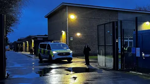 Modern brown brick school building seen in half dark with a police officer standing outside and a police van
