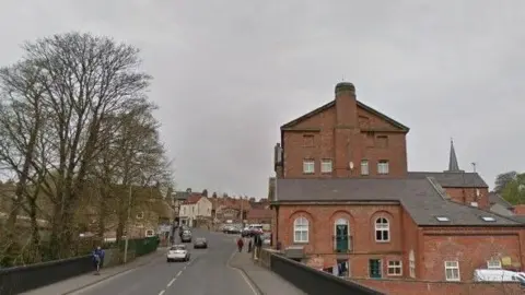 Google A road bridge with a large redbrick building on the right-hand side of the image, with a pavement and railings flanking the road. Cars and pedestrians are in the far distance.