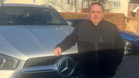 Supplied A man with a dark cardigan leaning against his silver car that has been parked on the driveway