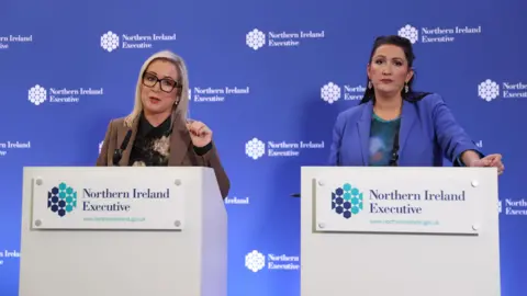PA Media A picture of Michelle O'Neill, a blonde haired woman with glasses and wearing a brown blazer, and Emma Little-Pengelly, a black haired woman wearing a blue blazer, standing beside each other at podiums at a press conference in front of a blue background.