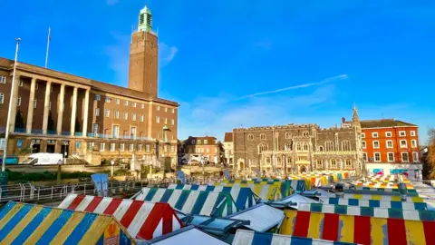 Rows of stalls at Norwich markets in Norwich city centre. The canopies are colourful and striped. In the background is the large imposing building of City Hall, with its tower furthest from the camera. There are blue skies above.