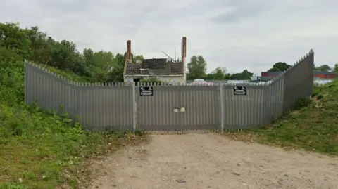 Google A fenced-off site with a locked metal gate and warning signs, with the remains of a fire-damaged house and chimney stacks visible behind it, surrounded by overgrown greenery.