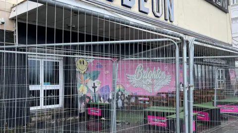 Scaffolding in the front of a complex which has a large pink grafitti-style mural which reads "Brighton". 