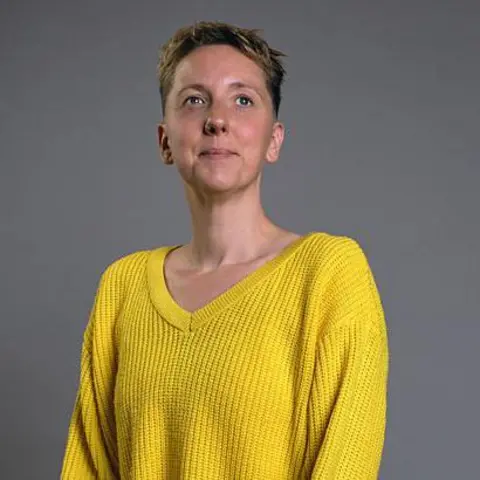 BBC Scotland A woman with short, cropped hair. She wearing a yellow V-necked jumper, and is sitting proud, looking into the distance.