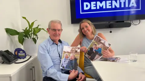 An older man sits with his daughter, holding up photos of their families 