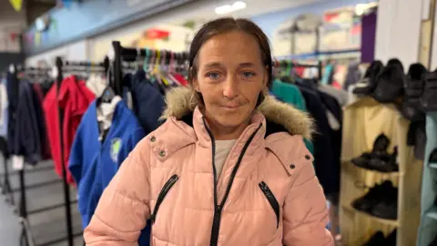 Rosemary Randall standing in the charity's school uniform store, smiling at the camera. She has brown hair tied back and is wearing a pink coat. She has a silver piercing above her lip.