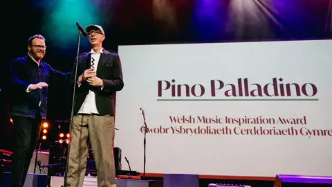 Welsh Music Prize Pino Palladino wearing a white shirt, black jacket and brown trousers. He is standing on the stage with Huw Stephens. Behind him there is a screen that says 'Pino Palladino, Welsh Music Inspiration Award'. It is translated in Welsh too. 