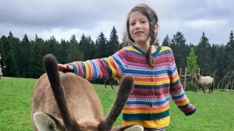 Aisha Cox smiles with a donkey. She is wearing a striped, multi coloured cardigan. In the background there is a forest lined with trees in a rural area.