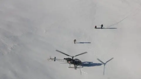 Reuters Una vista aérea de un helicóptero aterrizando en la nieve cerca de dos personas con una tercera persona caminando entre ellas.